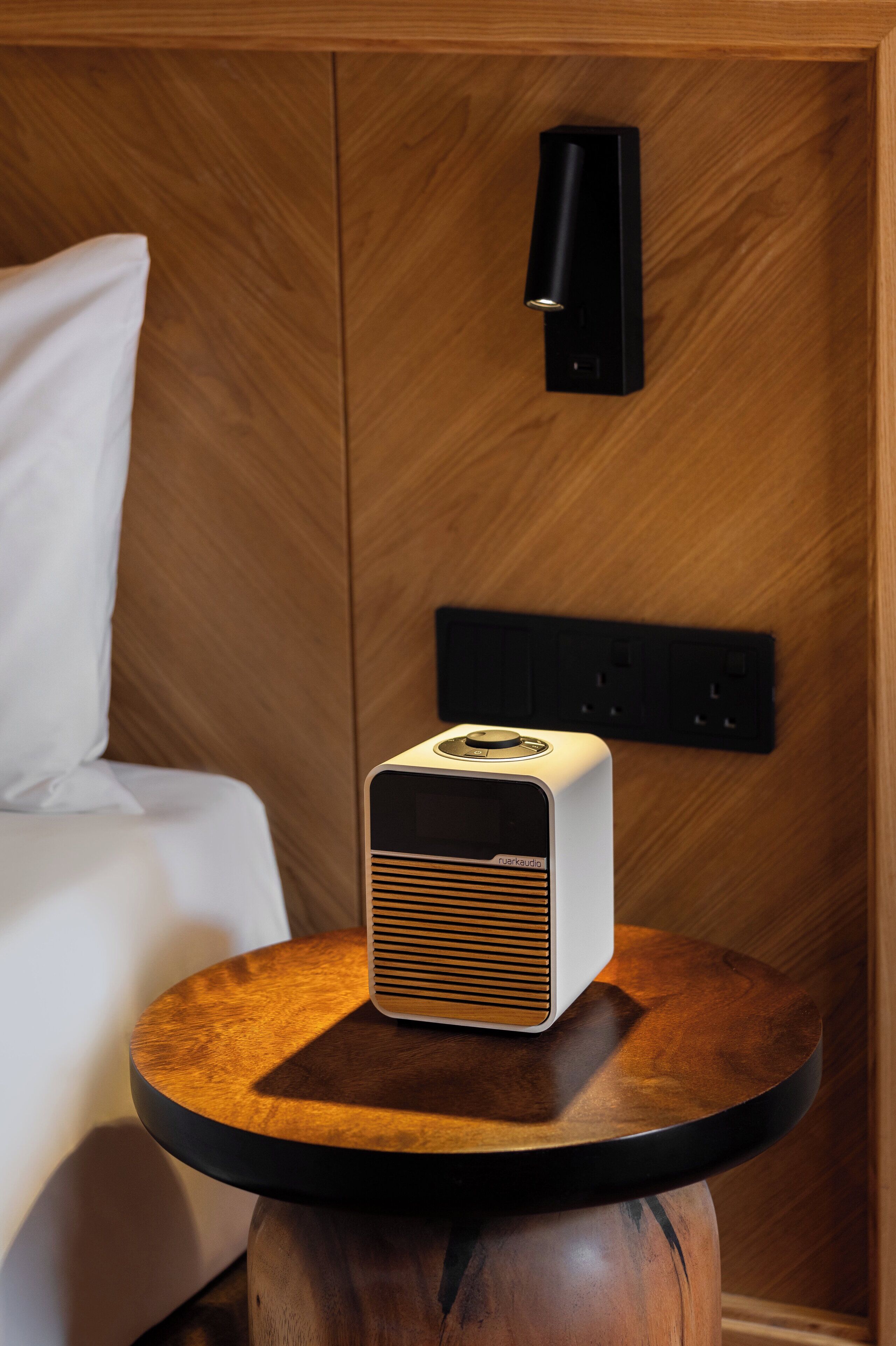 Una habitación moderna con un panel de pared de madera, una mesita de noche con un altavoz blanco y un aplique negro fijado a la pared.