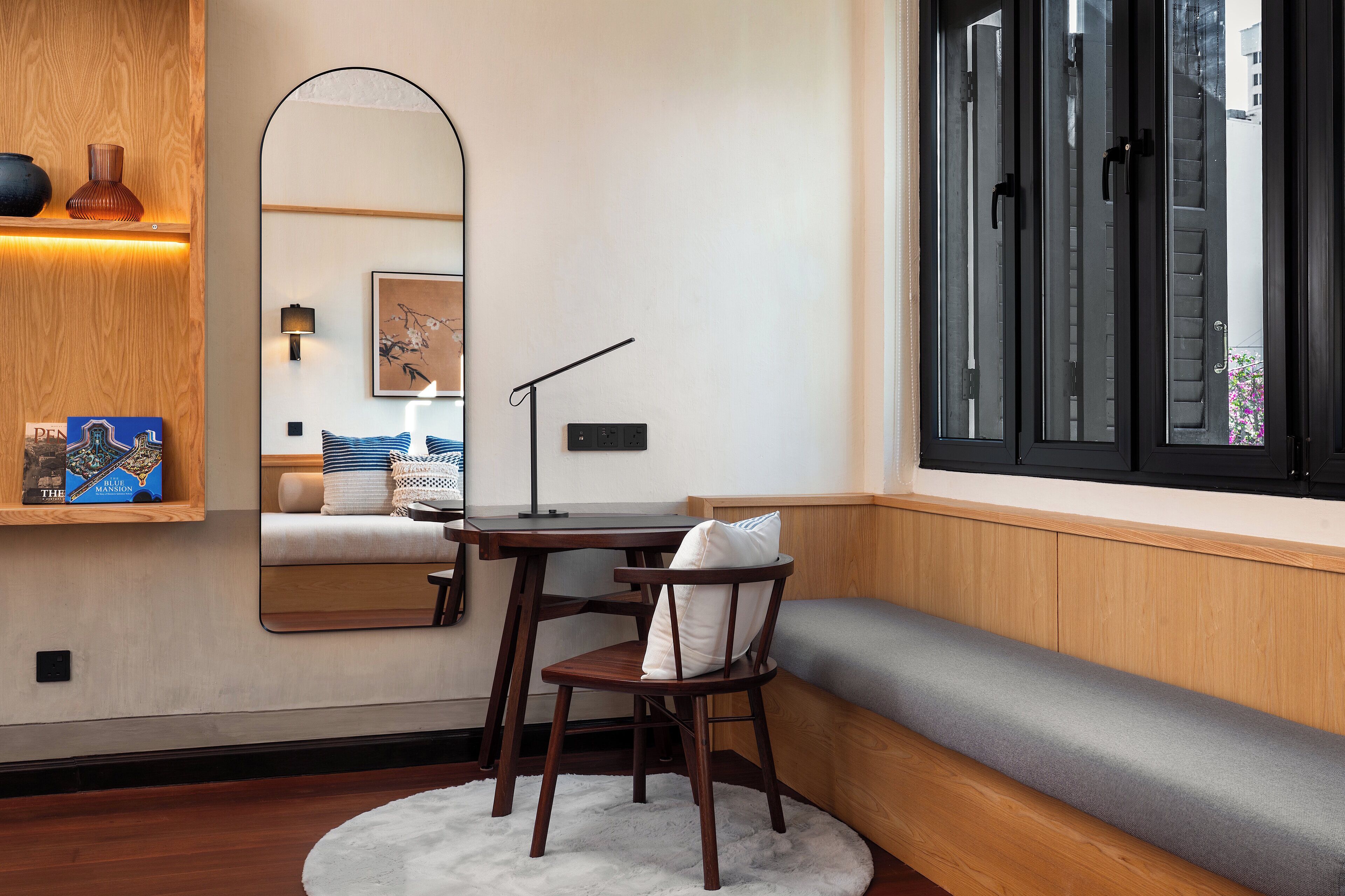 A corner room with a wooden bench, a desk with a chair, a mirror, and a window with blinds.