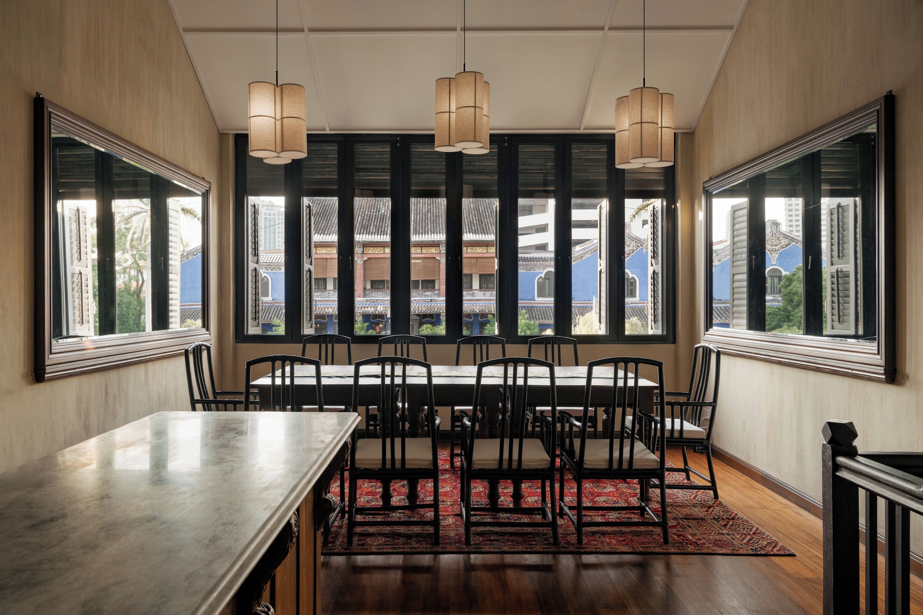 A dining area with a long table and chairs, large windows with a view of buildings, and hanging pendant lights.
