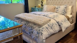 Een slaapkamer met een groot bed, een plafondventilator, een nachtkastje en een raam met gordijnen, en uitzicht op groen.