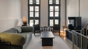 Ein modernes Wohnzimmer mit einer Couch, zwei Sesseln, einem Couchtisch, einem Fernseher und großen Fenstern mit Fensterläden.