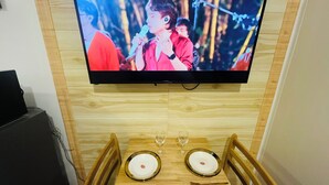 Uma mesa de jantar posta com pratos, talheres e copos de vinho, com uma televisão acima exibindo um artista.