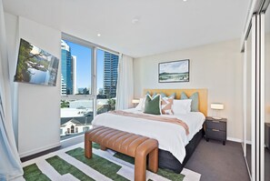 A hotel room with a large bed, a bench, a nightstand, and a view of tall buildings. - Hilton Residences - Pool Views (Surfers Paradise)