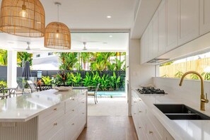 A modern kitchen with white cabinetry, a central island, and a view of a pool and tropical plants. - Baha Villa 89A | Luxury Holiday Home (Port Douglas)