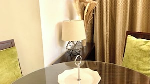 Mesa de vidro redonda com um prato branco e um castiçal de metal. Há duas cadeiras com almofadas verdes, um abajur com base estampada e uma cortina com padrão circular.