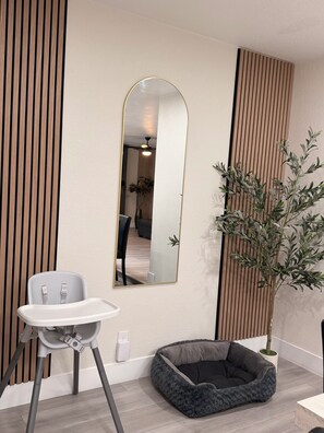 Ein modernes Zimmer mit einem hochgelehnten Sessel, einem kleinen Haustierbett, einem Blumentopf und einem an der Wand befestigten Spiegel.