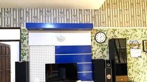 A living room with a TV on a white stand, a clock on the wall, and a wooden cabinet. - Homestay Jogja Dekat UII By Simply Homy (Ngaglik)