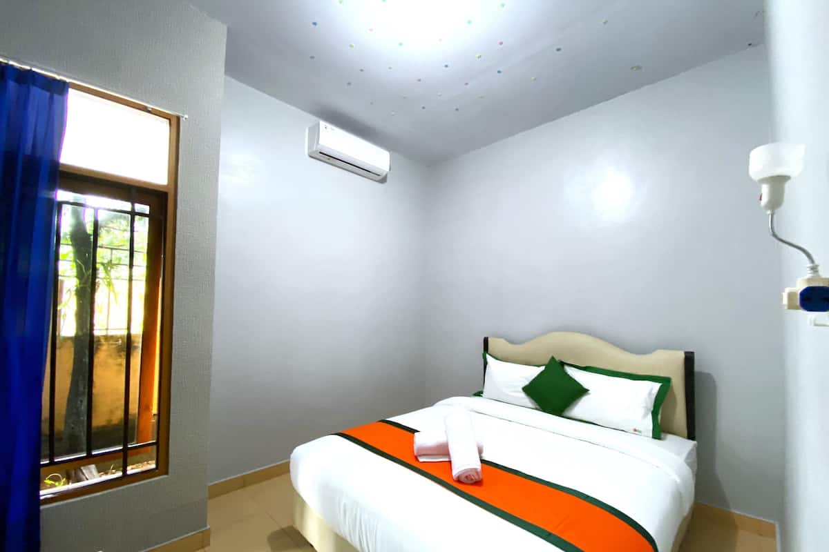 A hotel room with a bed, a window with blue curtains, an air conditioning unit, and a wall-mounted light fixture.