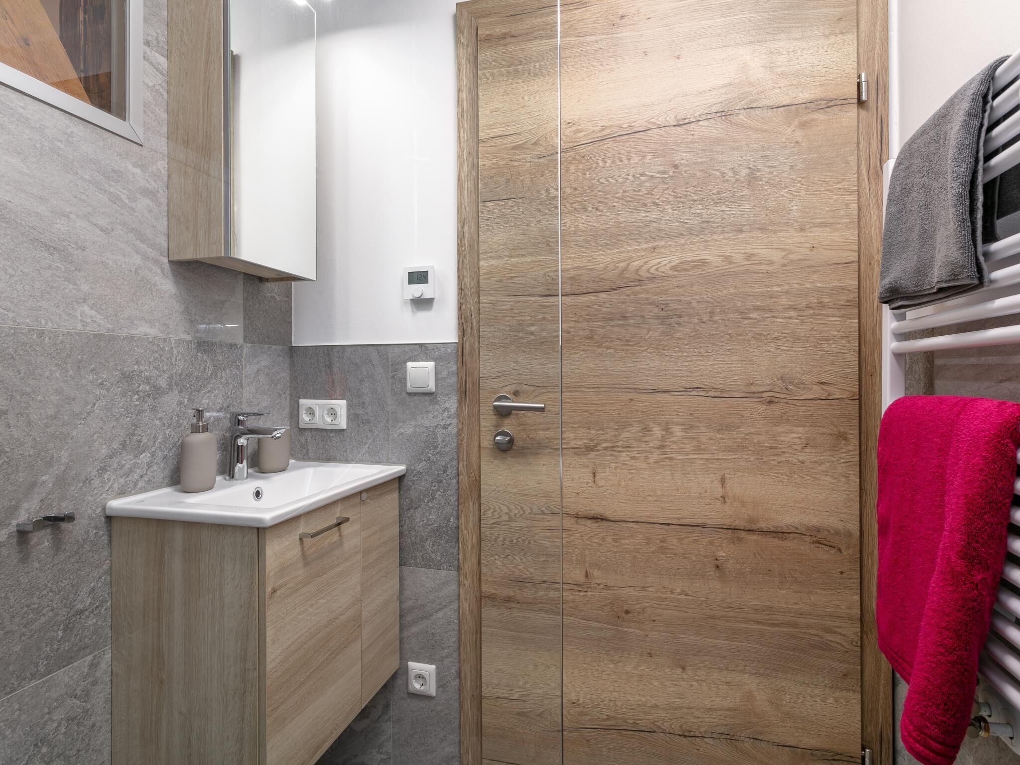 Casa de banho moderna com porta de madeira, uma pia com espelho e um porta-toalhas com uma toalha vermelha.
