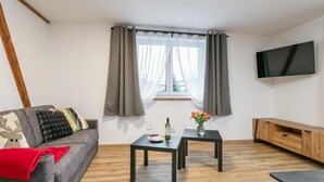 Ein modernes Wohnzimmer mit einem grauen Sofa, einem kleinen Tisch mit Blumen, einem Weinglas und einem flachbildfernseher an der Wand.
