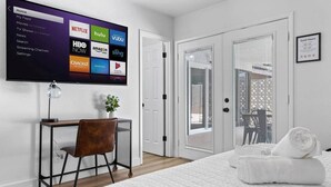 Et hotelværelse med et stort fjernsyn der viser streamingtjenester, et skrivebord med en stol, en seng med et sammenrullet håndklæde og en balkon med markiser.