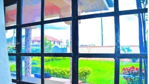 A room with a large window featuring a grid pattern, a tiled column, and a view of a green garden with a building in the background. - Majestic Airport Villa (Syokimau)