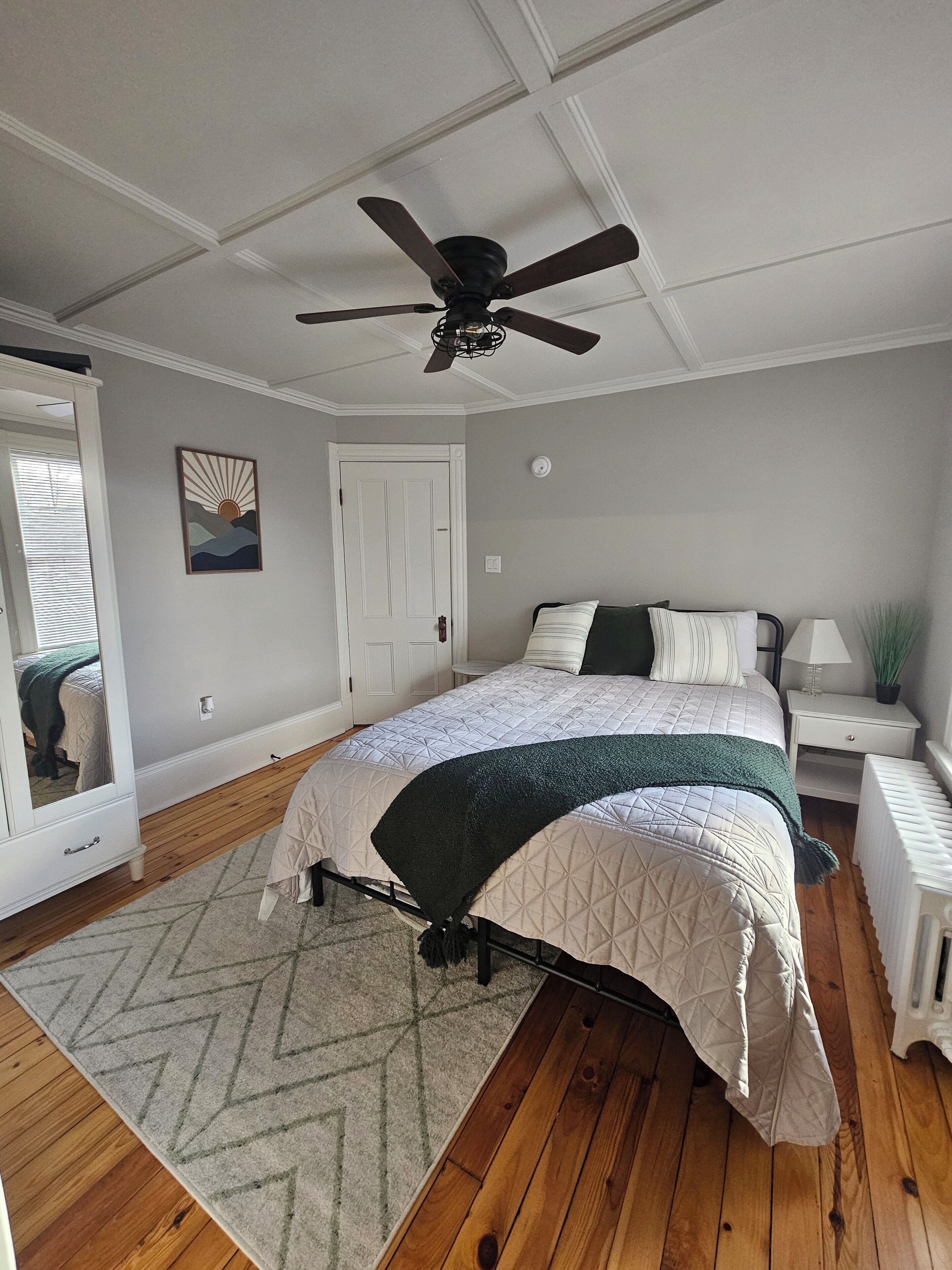 A bedroom with a bed, a ceiling fan, a nightstand, a mirror, and a door.