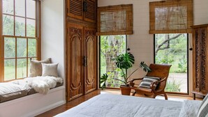 Une pièce comprenant un grand lit, une armoire en bois, un coin fenêtre avec des coussins, une chaise en bois et une plante en pot.