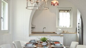 Un coin repas avec une table en bois dressée pour quatre personnes, des chaises en osier blanches, un lustre et, au fond, une cuisine.