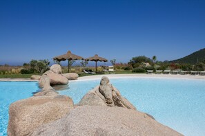 Een helderblauw zwembad met grote rotsen op de voorgrond, een zandstrand en rieten parasols op de achtergrond.
