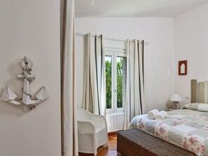 Une chambre à coucher comprenant un lit, un banc en osier, une table de chevet avec une lampe, une fenêtre avec des rideaux et un élément décoratif en forme d’ancre fixé au mur.