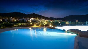 A swimming pool at twilight with surrounding buildings and hills. - Li Conchi 35 – Spacious Holiday Home for 7 in a Modern Resort (Castiadas)