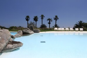 Piscina azul cristalina con sillas reclinables blancas y palmeras al fondo.