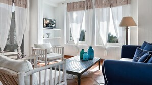 A room with a white chair, a blue sofa, a wooden table, and a lamp. - Li Conchi 35 – Spacious Holiday Home for 7 in a Modern Resort (Castiadas)