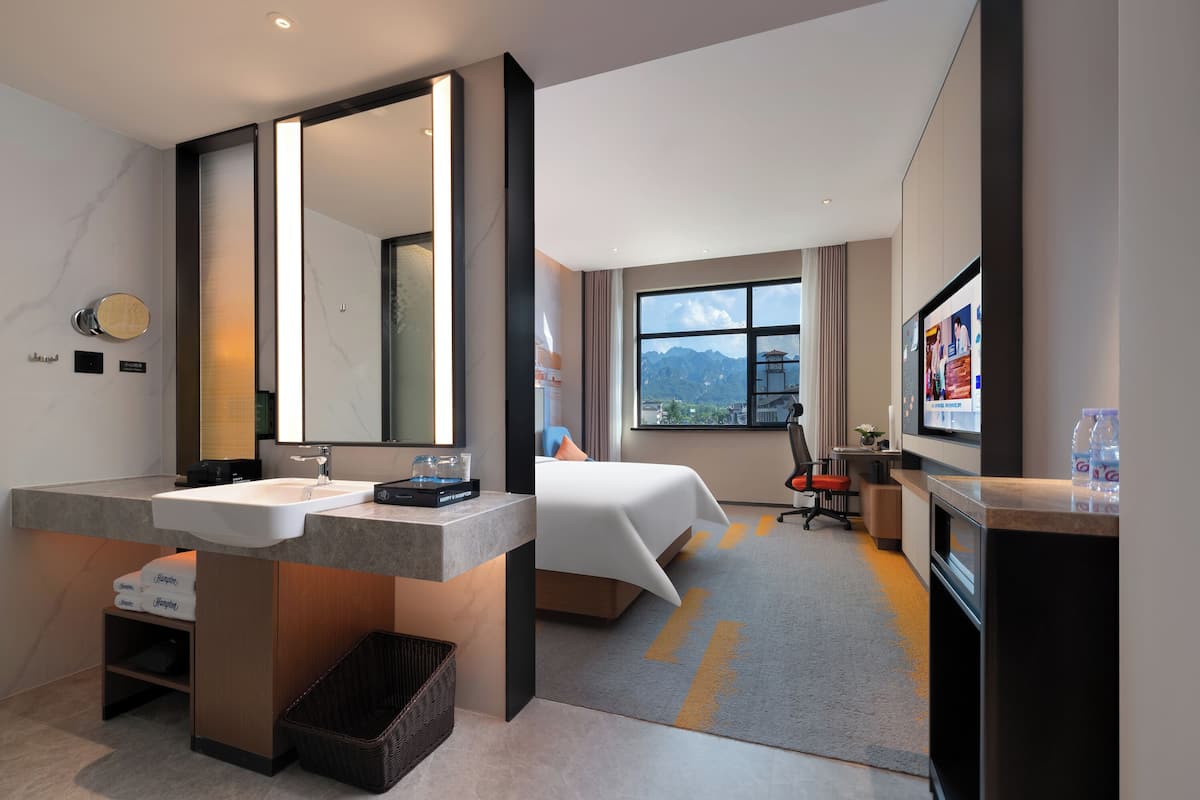 Ein Hotelzimmer mit einem großen Spiegel, einem Bett, einem Schreibtisch, einem Sessel und einem Fernseher, mit Blick auf Berge.