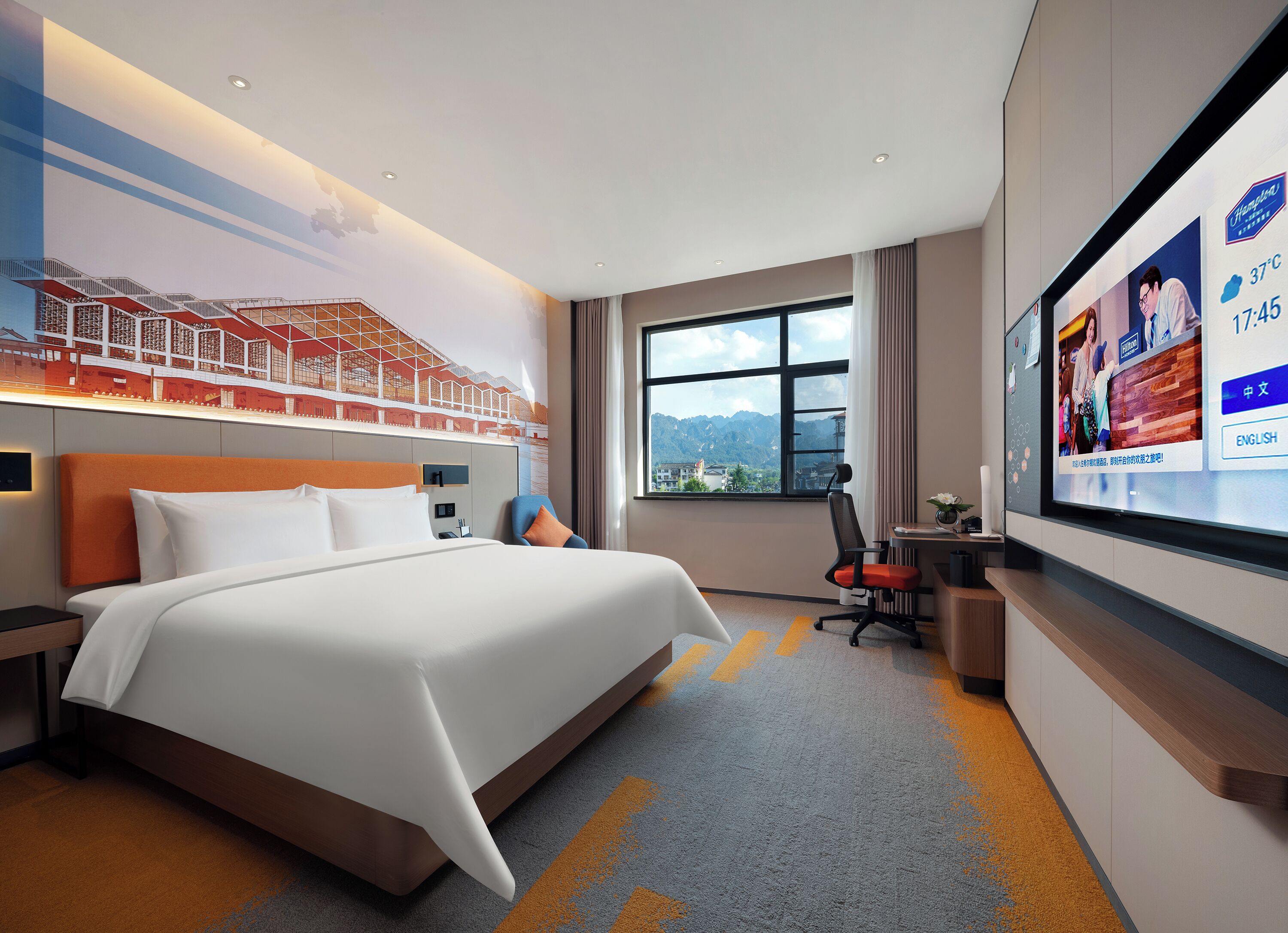 A hotel room with a large bed, a desk with a chair, a TV on the wall, and a window with a mountain view.