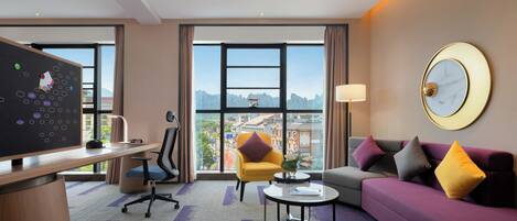 A modern hotel room with a large desk, a comfortable sofa, a small coffee table, and a view of the cityscape through large windows.