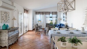 Un comedor con una mesa lista para cuatro personas, sillas de mimbre, una araña y vistas a través del ventanal.