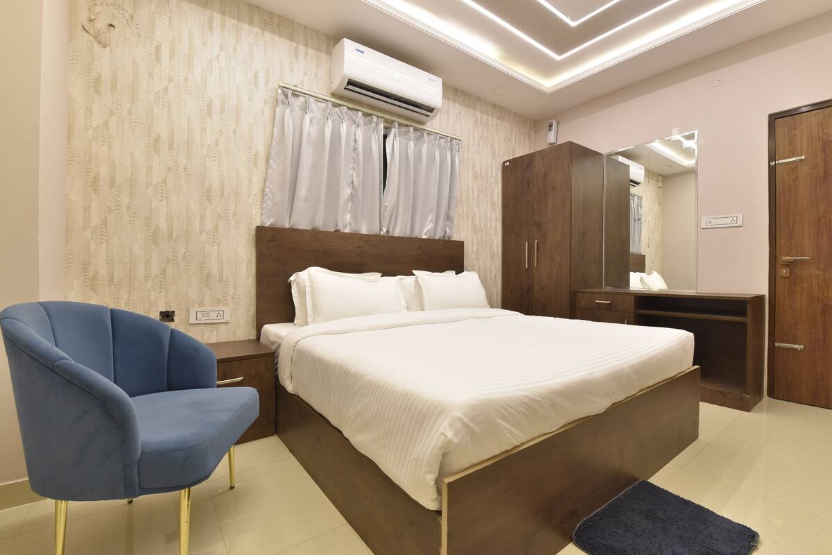 A bedroom with a bed, a blue chair, a wooden wardrobe, and an air conditioning unit.