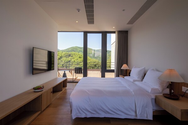 A modern hotel room with a large bed, a TV, a desk with a bowl of fruit, and a balcony with a view of green hills. - LOSTVILLA Green Gable-Anji (Huzhou)