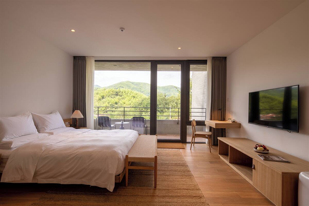 Habitación de hotel moderna con una cama grande, un escritorio, un televisor y un balcón con vistas a colinas verdes.