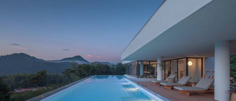 Una piscina infinita moderna con terraza de madera y sillones de descanso, con vista a un paisaje montañoso.