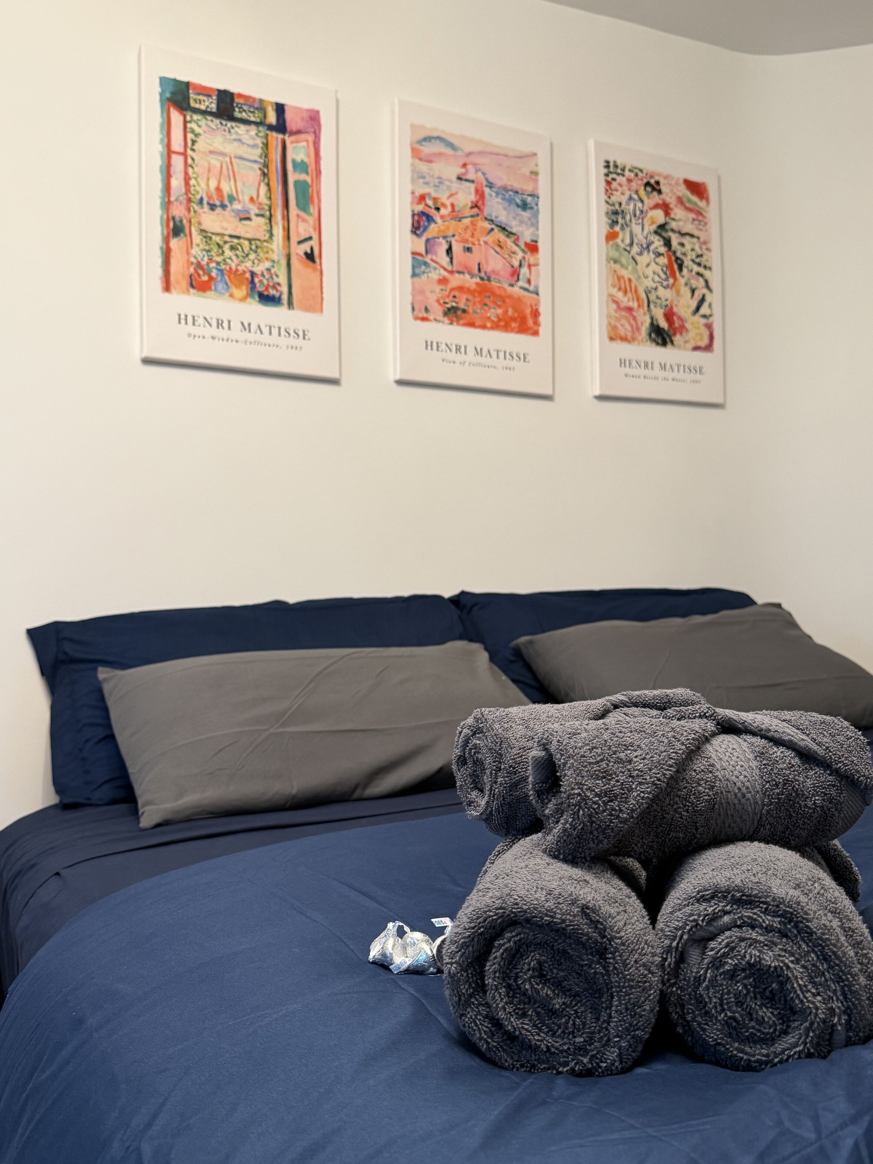 Una cama con cubrecama azul, almohadas grises y una pila de toallas grises. Tres obras enmarcadas de Henri Matisse están exhibidas en la pared sobre la cama.