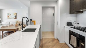 Une cuisine moderne avec des comptoirs blancs, un robinet noir et une cuisinière. On y trouve un coin repas avec une table et des chaises en bois, ainsi qu'un mur blanc orné d'un tableau.