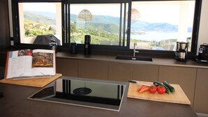 A modern kitchen with a built-in stove, a wooden cutting board with vegetables, a coffee maker, and a view of a lake and mountains through large windows. - Villa d'exception piscine privée 🏝️ 300m² - 16 pers Promo en cours (Pietrosella)