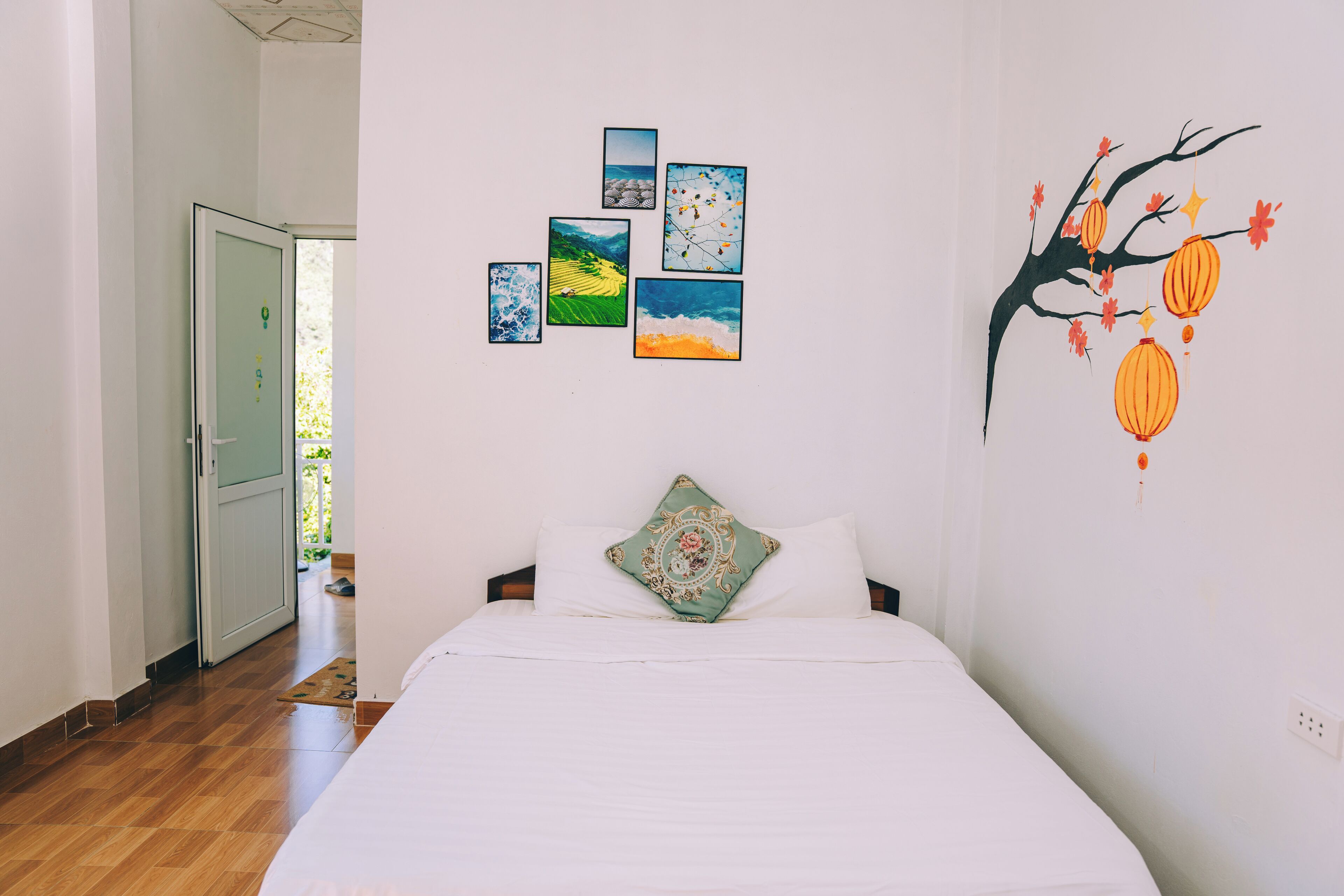 A bedroom with a bed, a door, and wall art including a tree with lanterns.