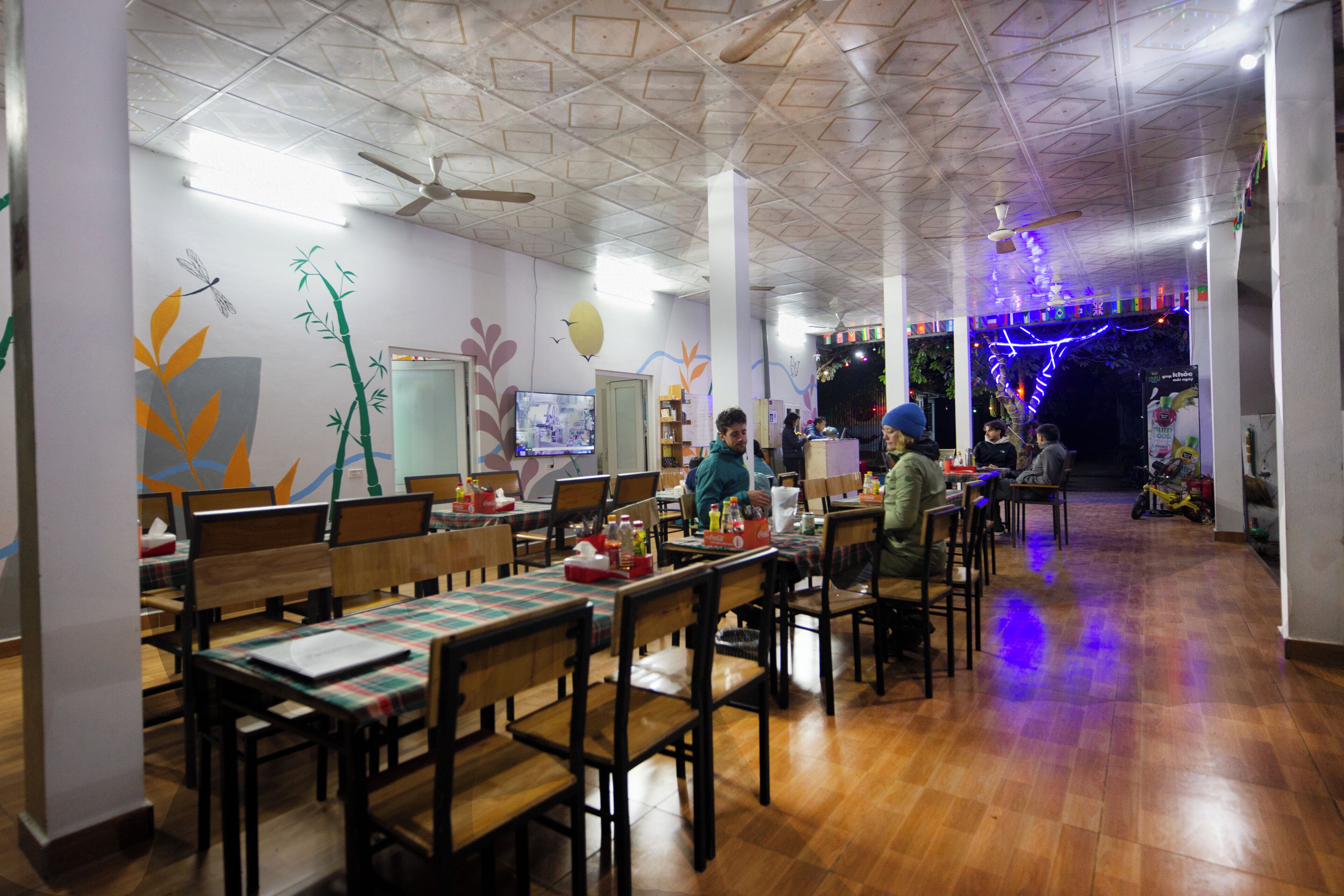 Restaurante com mesas e cadeiras de madeira, um ventilador de teto e decorações nas paredes, incluindo uma libélula e bambu.