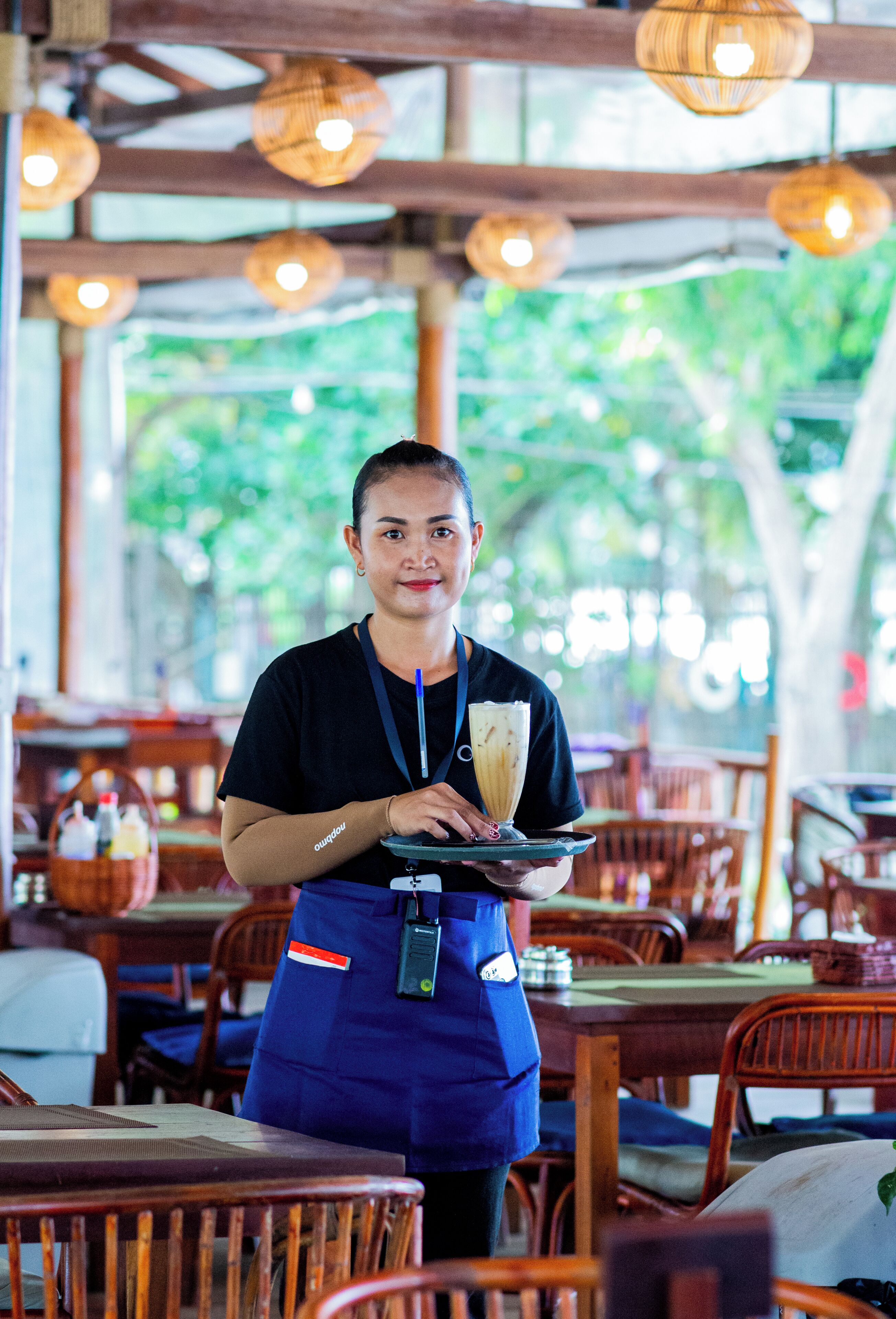 A person in a blue uniform holding a tray with a beverage, standing in a restaurant with wooden furniture and hanging lights.