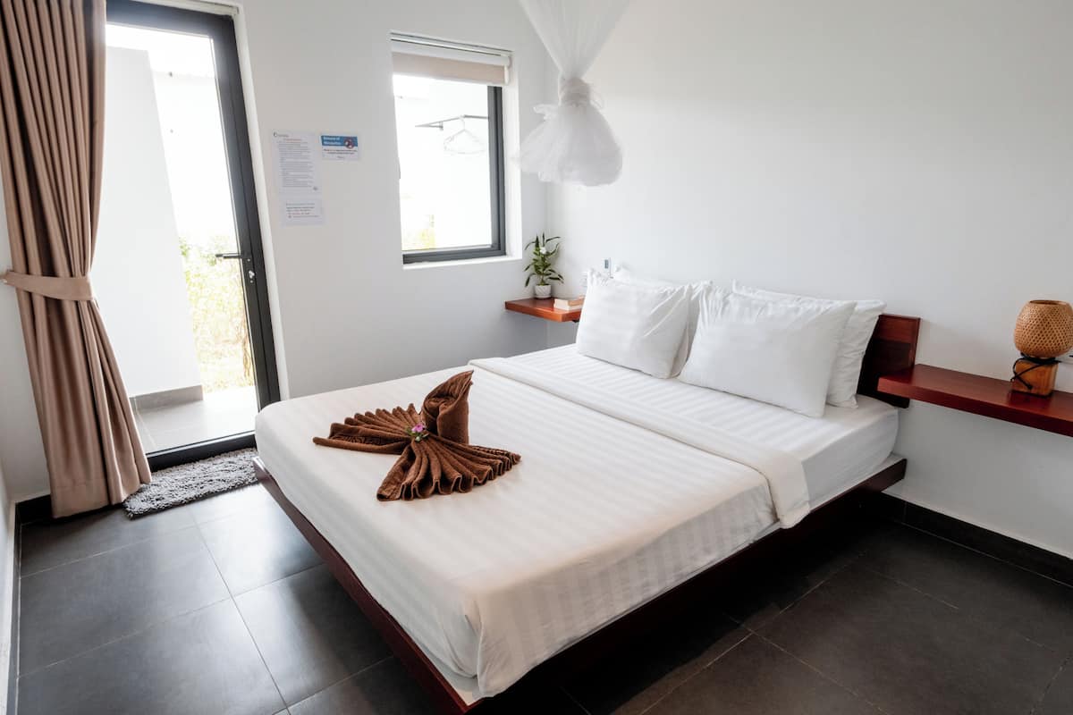 Una habitación de hotel con cama, ventana, mesita de noche y cortina.