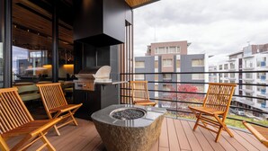 Een balkon met een vuurkorf van steen, houten stoelen en een barbecue.
