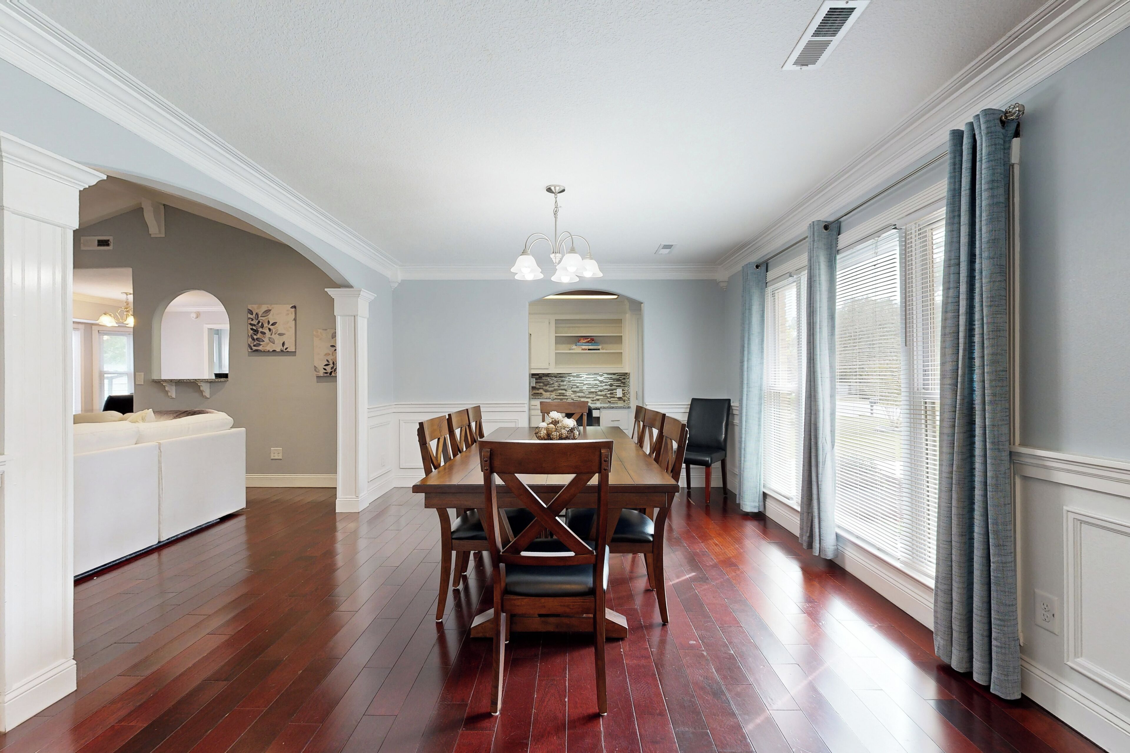 A dining area with a wooden table and chairs, a white sofa, and a kitchen in the background.
