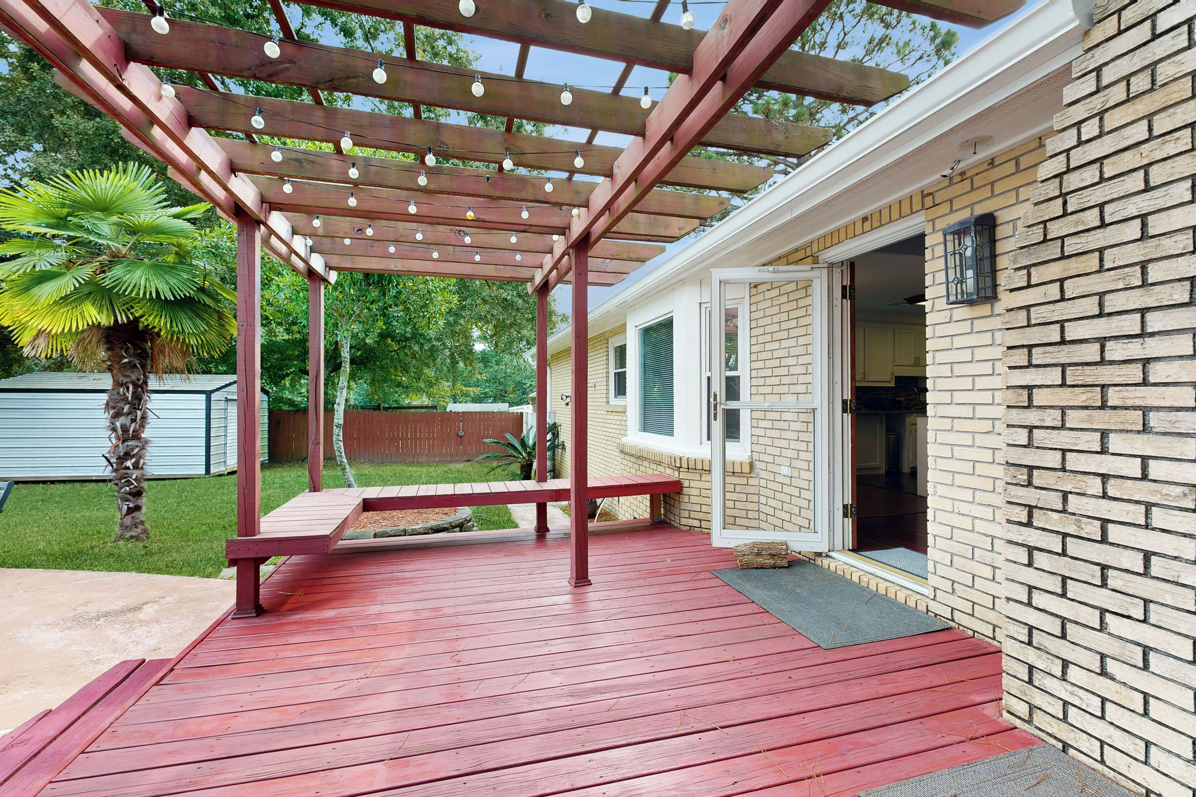 A covered patio with a wooden pergola and string lights, a palm tree, and a brick house with a door open.