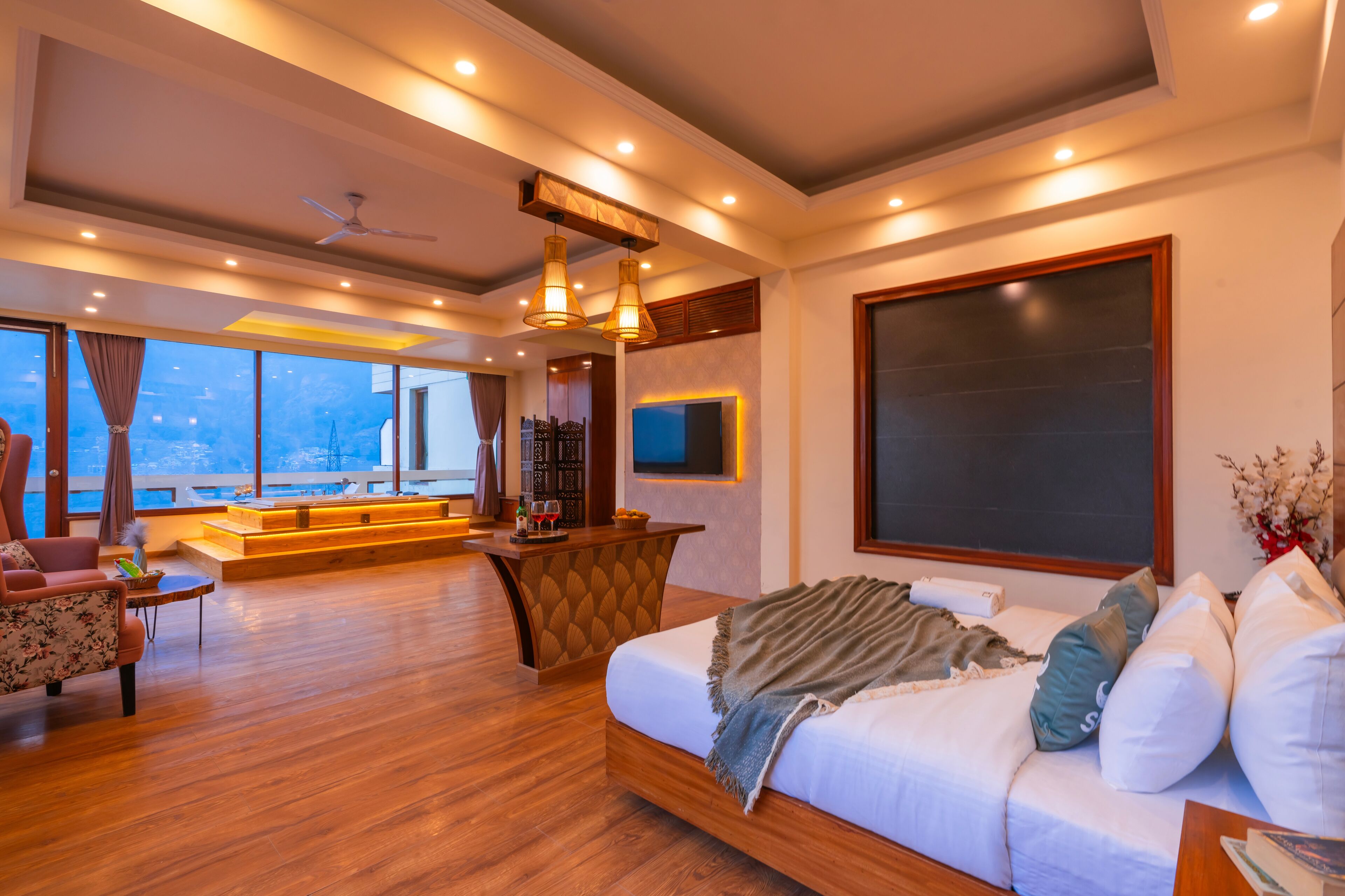 Ein großzügiger Wohnbereich mit einem großen Bett, Holzböden, einer Sitzecke mit Sofa und Sesseln, einem an der Wand montierten Fernseher und Blick ins Freie durch große Fenster.
