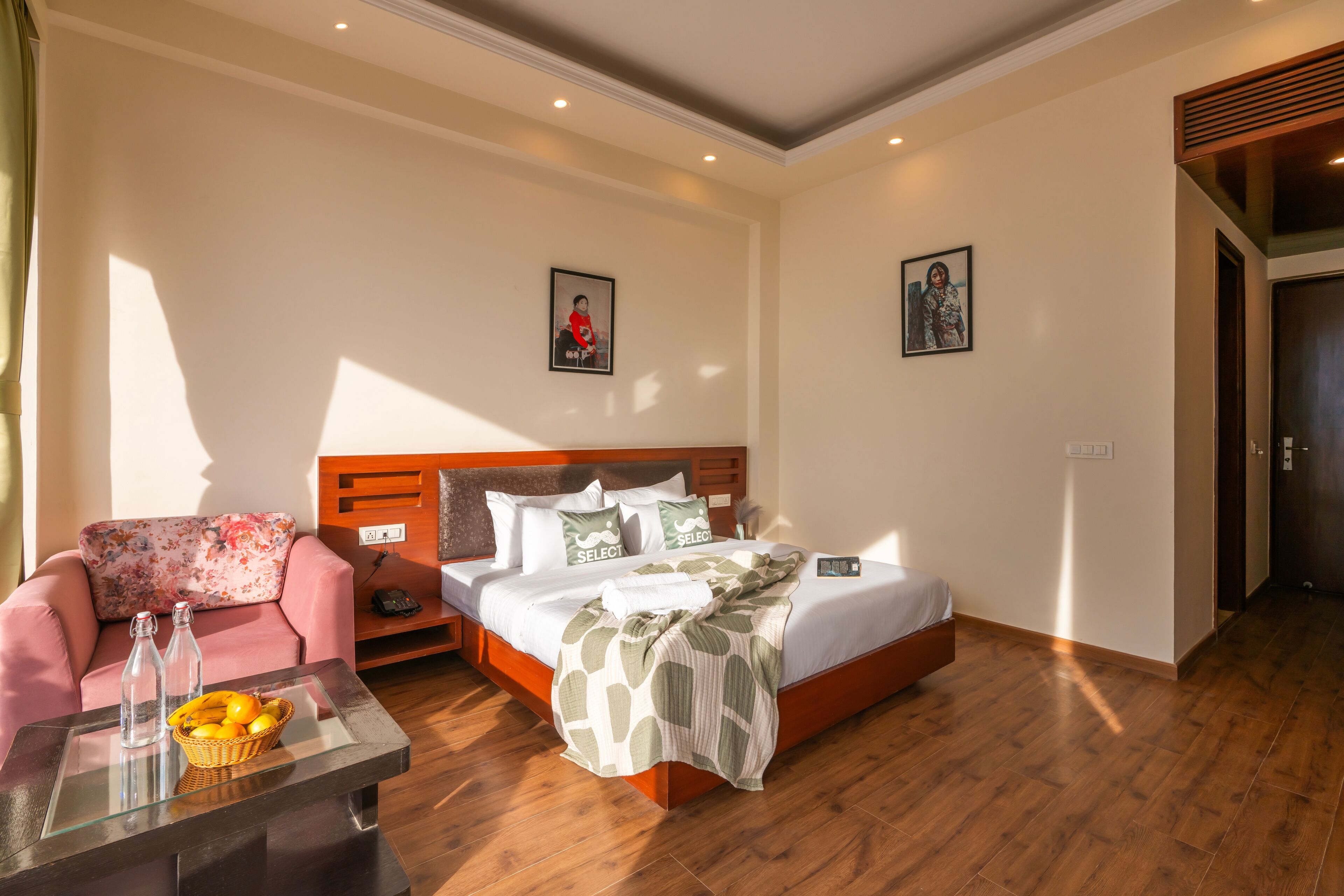 A hotel room with a bed, a sofa, a coffee table with bottles and fruit, and two framed pictures on the wall.