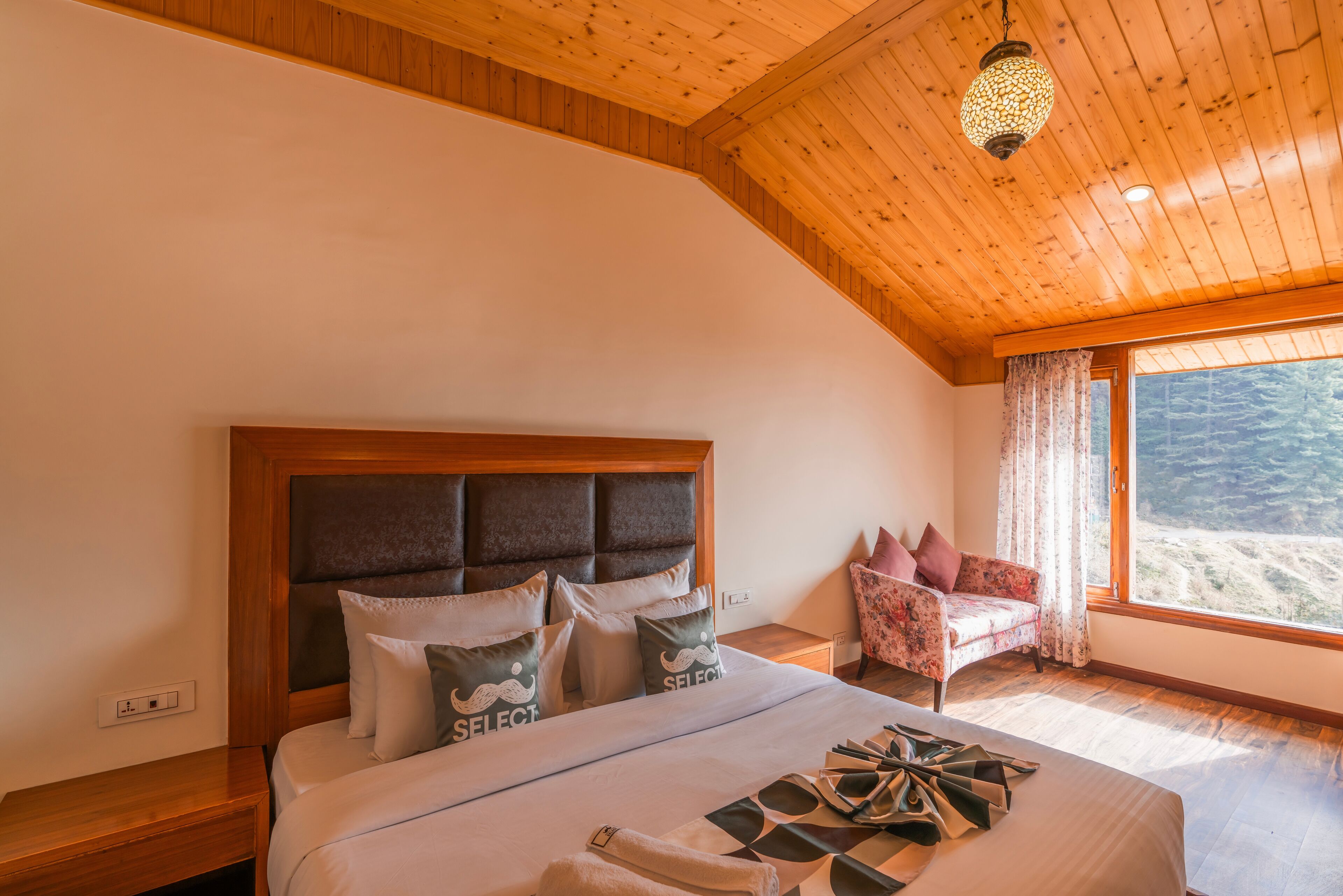 A bedroom with a wooden ceiling, a large bed with multiple pillows, a nightstand, a chair, and a window with a view of the outdoors.