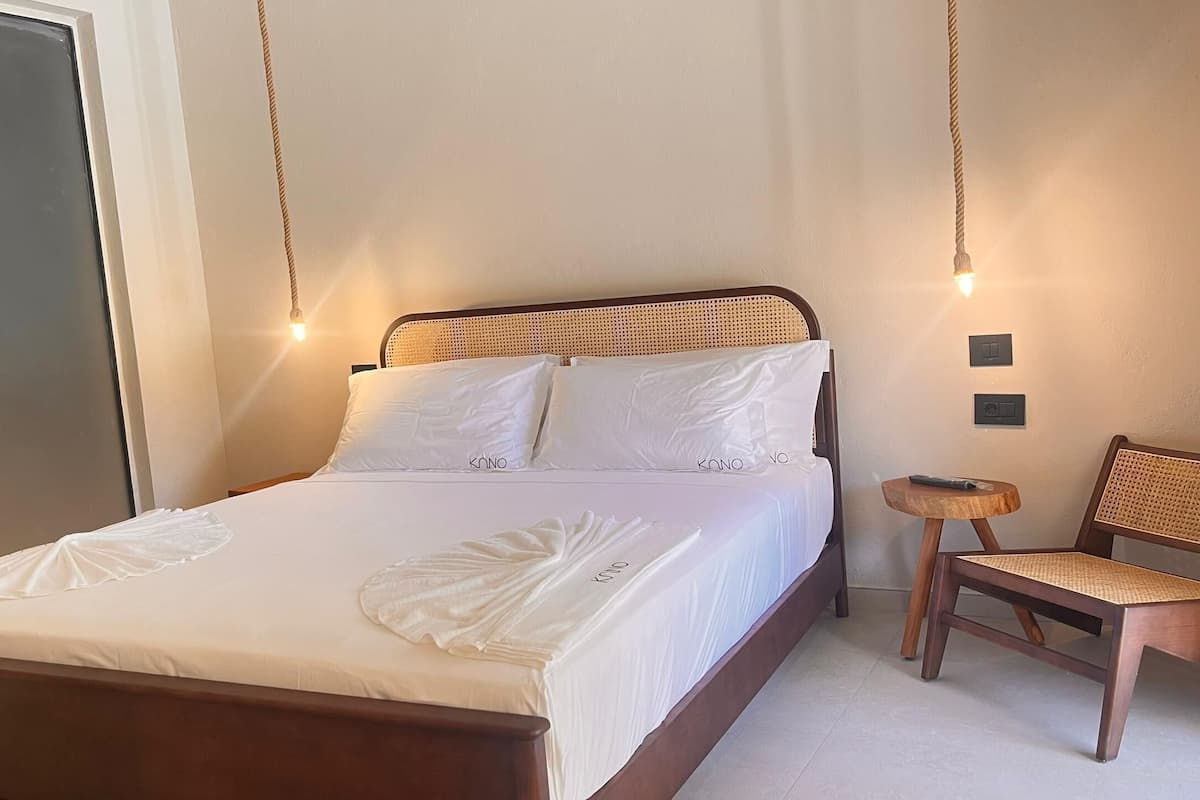 Una cama bien hecha con sábanas y almohadas blancas, un cabecero de madera, una pequeña silla de madera con asiento de mimbre y una lámpara de pared.