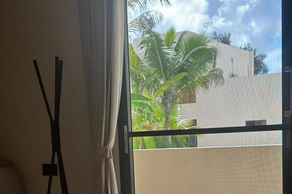 Habitación de hotel con cama, toallas blancas, ventana con vista a palmeras y edificios.
