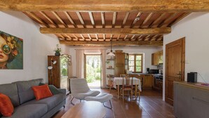 Ein gemütliches Wohnzimmer mit einer Couch, Sesseln, einem Holztisch und einer Küchenzeile mit Holzausbau und Fliesenboden.
