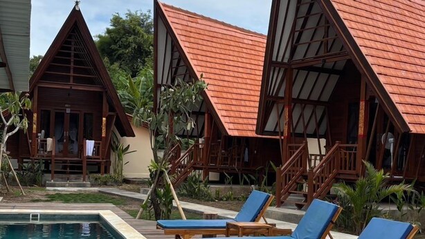 Un área junto a la piscina con cubierta de madera, sillones de descanso y bungalows sobre el agua con tejados rojos.