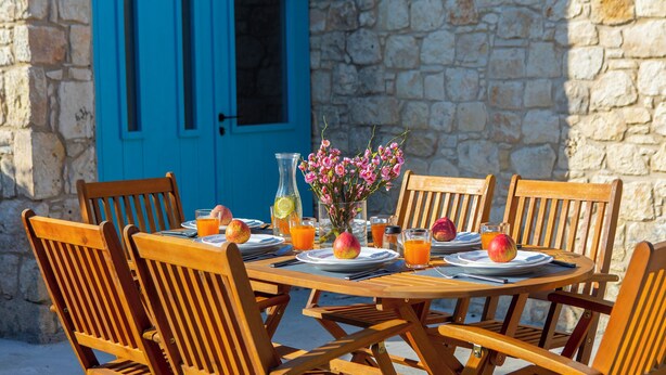 A wooden table set for breakfast with chairs, a vase of flowers, and a blue door in the background. - AKAMANTIDA PLATEA 2. Country house of the 1900s. Restored to its authentic charm (Pano Arodes)